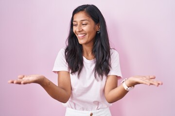 Young hispanic woman standing over pink background smiling showing both hands open palms, presenting and advertising comparison and balance