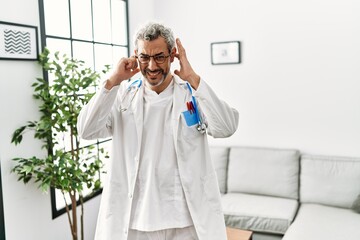 Wall Mural - Middle age hispanic man wearing doctor uniform and stethoscope at waiting room covering ears with fingers with annoyed expression for the noise of loud music. deaf concept.