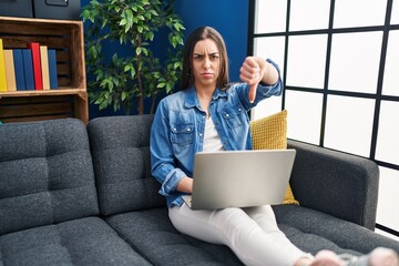 Sticker - Hispanic woman using laptop at home with angry face, negative sign showing dislike with thumbs down, rejection concept