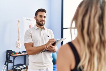 Poster - Young hispanic man wearing physiotherapist uniform writing on clipboard at clinic