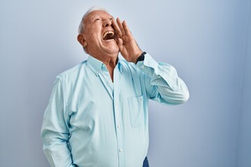 Canvas Print - Senior man with grey hair standing over blue background shouting and screaming loud to side with hand on mouth. communication concept.