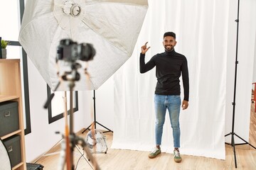 Canvas Print - Young hispanic man with beard posing as model at photography studio smiling and confident gesturing with hand doing small size sign with fingers looking and the camera. measure concept.