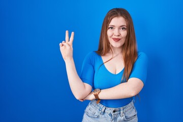 Wall Mural - Redhead woman standing over blue background smiling with happy face winking at the camera doing victory sign with fingers. number two.