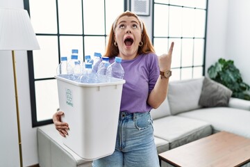 Sticker - Young redhead woman holding recycling wastebasket with plastic bottles amazed and surprised looking up and pointing with fingers and raised arms.