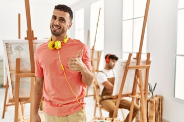 Poster - Young hispanic man at art studio doing happy thumbs up gesture with hand. approving expression looking at the camera showing success.