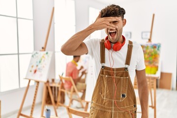 Poster - Young hispanic man at art studio peeking in shock covering face and eyes with hand, looking through fingers with embarrassed expression.