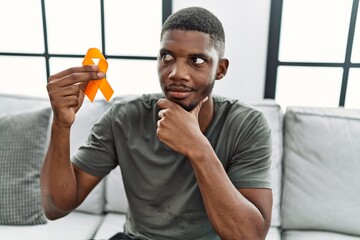 Sticker - Young african american man holding awareness orange ribbon sitting on the sofa serious face thinking about question with hand on chin, thoughtful about confusing idea