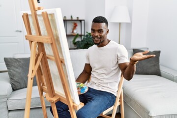 Sticker - Young african man painting on canvas at home smiling cheerful presenting and pointing with palm of hand looking at the camera.