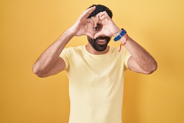 Sticker - Hispanic man with beard standing over yellow background doing heart shape with hand and fingers smiling looking through sign