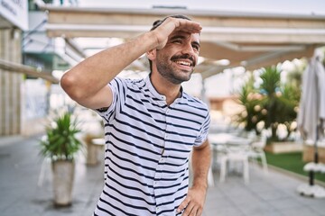 Wall Mural - Young hispanic man with beard outdoors at the city very happy and smiling looking far away with hand over head. searching concept.