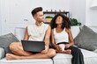 © Krakenimages.com - Young interracial couple using laptop at home sitting on the sofa smiling looking to the side and staring away thinking.