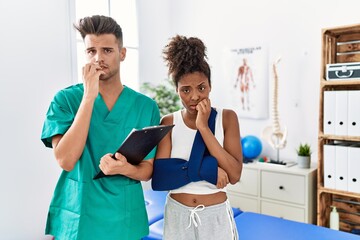 Canvas Print - Physiotherapist working with patient wearing arm on sling at rehabilitation clinic looking stressed and nervous with hands on mouth biting nails. anxiety problem.