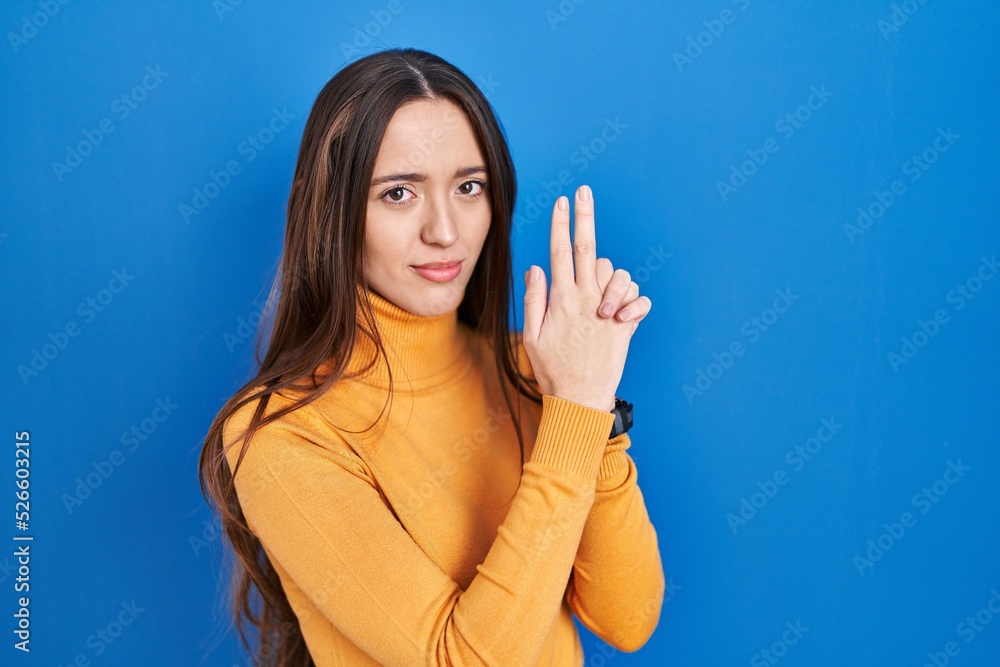 Young brunette woman standing over blue background holding symbolic gun with hand gesture, playing killing shooting weapons, angry face