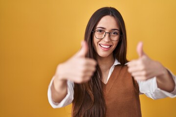 Wall Mural - Young brunette woman standing over yellow background wearing glasses approving doing positive gesture with hand, thumbs up smiling and happy for success. winner gesture.