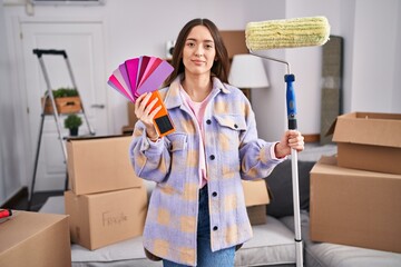 Sticker - Young brunette woman painting new home walls with paint roller relaxed with serious expression on face. simple and natural looking at the camera.