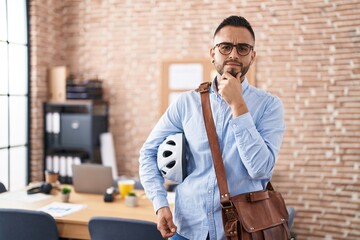 Sticker - Young hispanic man working at the office holding bike helmet serious face thinking about question with hand on chin, thoughtful about confusing idea