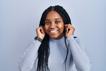 Wall Mural - African american woman standing over blue background smiling pulling ears with fingers, funny gesture. audition problem