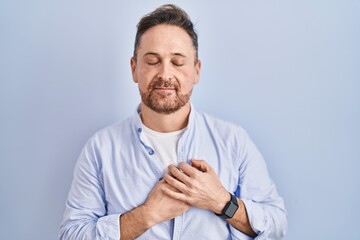 Sticker - Middle age caucasian man standing over blue background smiling with hands on chest with closed eyes and grateful gesture on face. health concept.