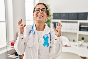 Wall Mural - Young brunette doctor woman wearing stethoscope at the clinic gesturing finger crossed smiling with hope and eyes closed. luck and superstitious concept.