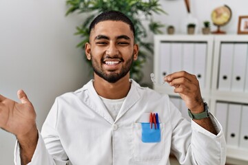 Wall Mural - Young indian man working at dentist clinic holding invisible aligner celebrating achievement with happy smile and winner expression with raised hand