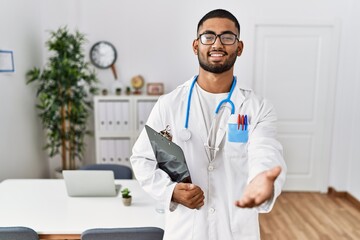 Sticker - Young indian man wearing doctor uniform and stethoscope smiling cheerful offering palm hand giving assistance and acceptance.