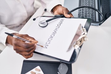 Poster - African american woman wearing doctor uniform writing hypothyroidism on clipboard at clinic