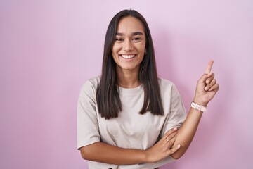 Wall Mural - Young hispanic woman standing over pink background with a big smile on face, pointing with hand finger to the side looking at the camera.