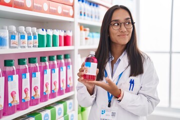 Poster - Young hispanic woman working at pharmacy drugstore holding syrup smiling looking to the side and staring away thinking.