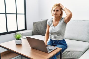 Middle age blonde woman using laptop at home confuse and wondering about question. uncertain with doubt, thinking with hand on head. pensive concept.
