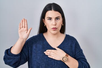 Canvas Print - Young brunette woman standing over isolated background swearing with hand on chest and open palm, making a loyalty promise oath