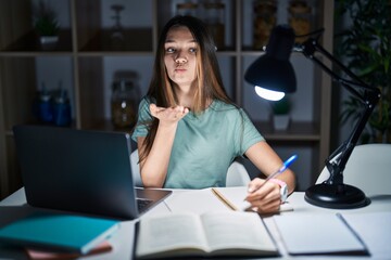 Sticker - Teenager girl doing homework at home late at night looking at the camera blowing a kiss with hand on air being lovely and sexy. love expression.