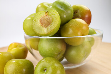 Wall Mural - Small mini green fresh juicy plum fruit in transparent glass bowl wooden table white background