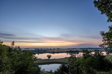 Scenic view of a reflective river flowing through the field with green trees at sunrise