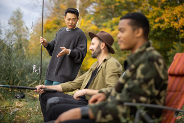 Asian man talking to male multiracial friends during fishing in nature. Men resting and spending time together on river or lake shore. Concept of leisure, hobby and weekend in nature. Friendship