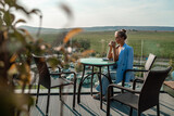 Fototapeta Uliczki - A middle-aged woman sits in a street cafe overlooking the mountains at sunset. She is dressed in a blue jacket and drinks coffee admiring the nature. Travel and vacation concept.