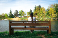 Young Woman Sitting On Bench In Front Of Lake At Park