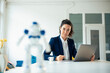 © Westend61 - Serious businesswoman sitting with laptop looking at toy robot on table