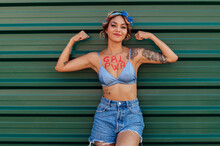 Woman With Girl Power Text On Chest Showing Biceps In Front Of Green Metal Wall