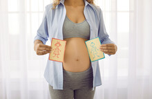 Two Cards With Picture Of Funny Boy And Girl That Woman Holds Near Her Pregnant Belly. Cropped Image Of Woman With Naked Pregnant Belly On Window Background With Cards For Gender Party. Middle Section