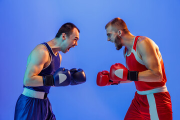 Sports emotions. Excited professional boxers in red and blue sports uniform boxing isolated on blue background in neon light. Sport, skills, power, training, energy