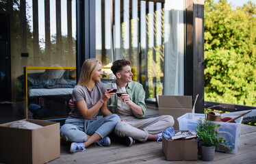 Cheerful young couple moving in their new tiny house in woods sitting on floor on terrace and drinking wine. Conception of moving and sustainable living.