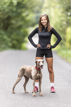 Young Sporting Woman And Her Weimaraner Dog Stand Together In The Forest