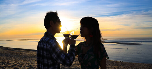 Wall Mural - Silhouette romantic couple  toasting each other with wine while sitting together on a sandy beach at sunset scene mood