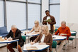 © Seventyfour - Group of senior students sitting at desks and making notes while teacher reading a lecture in classroom