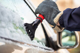 Fototapeta Zwierzęta - Hand in glove holding a snow scraper and removing ice from a car window with rear view mirror in background. Concept of danger on roads in winter season. Maintenance of vehicle in cold temperatures.