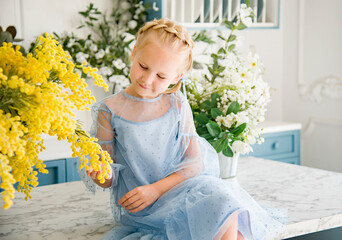 little smiing blond cute girl  in blue dress sitting in blue and white kitchen with yellow flowers mimosa 