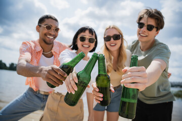 Wall Mural - interracial friends in sunglasses clinking beer bottles on blurred background.