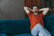 Young minded man wears red t-shirt hold hands behind neck look aside sit on blue sofa stay at home hotel flat rest relax spend free spare time in living room indoors grey wall. People lounge concept.