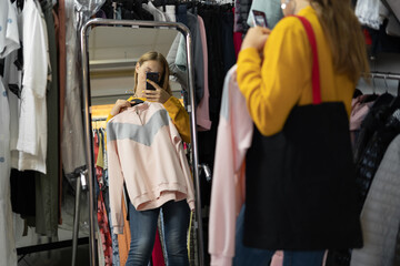 shopping, fashion, style and people concept - happy woman choosing clothes and looking to mirror in mall or clothing store