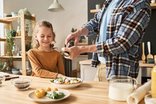 Close-up Of Young Dad Using Pepper Shaker To Add Spices In Vegetable Salad, He Preparing Together With His Son In Kitchen